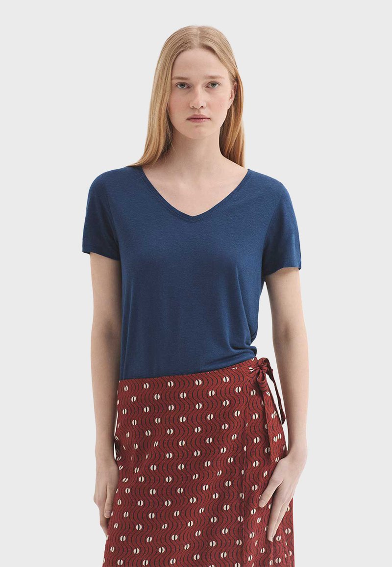 Jeune femme aux cheveux blonds raides portant un haut bleu marine à manches courtes avec un col en V et une jupe portefeuille rouge à motifs.