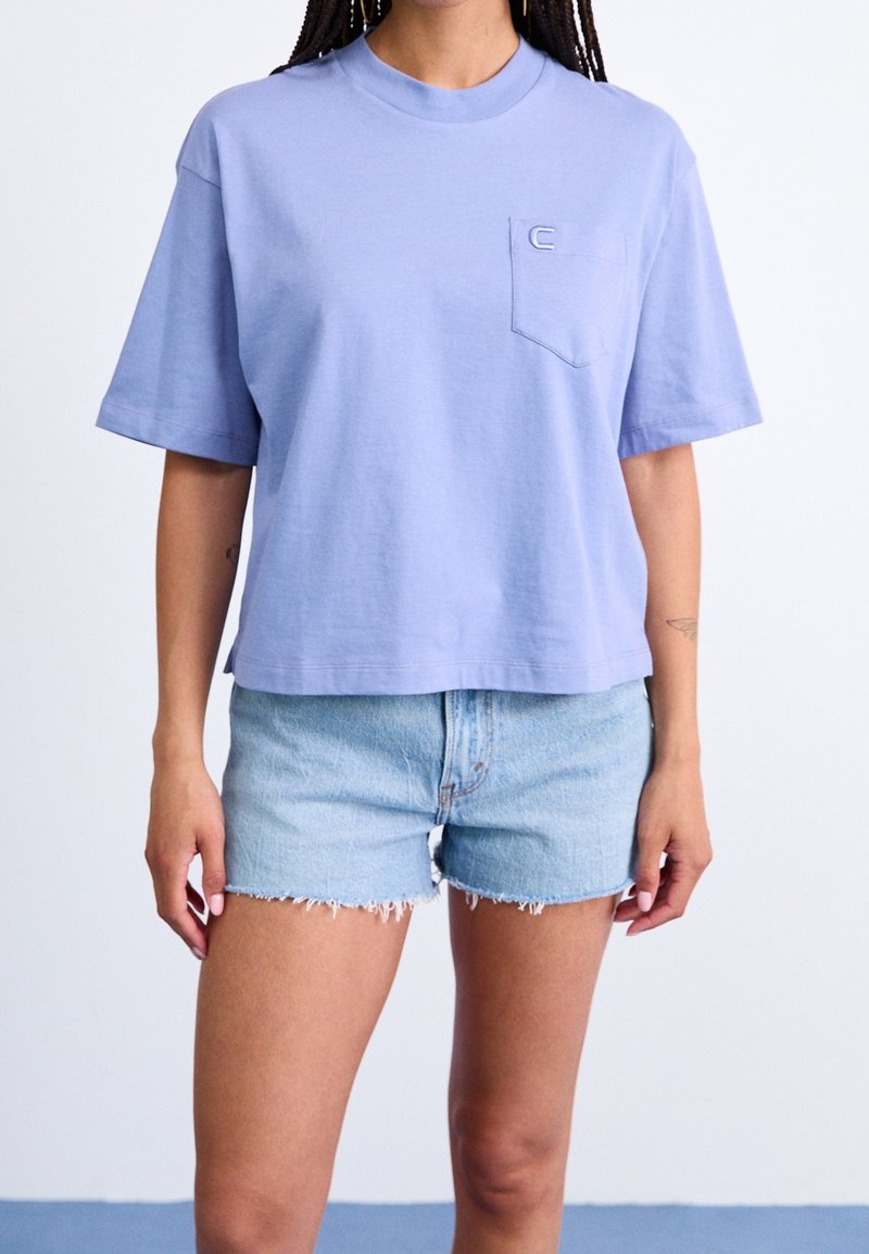 T-shirt in cotone color lilla chiaro con maniche corte, scollo rotondo e taschino sul petto a sinistra. Abbinata a pantaloncini di jeans sfrangiati.