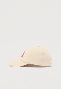 Μπεζ βαμβακερό καπέλο με κυρτό γείσο, διακοσμημένο με κόκκινο κεντητό σχέδιο στη μία πλευρά και τρύπες αερισμού στο πάνω μέρος. Ρυθμιζόμενο λουράκι στο πίσω μέρος.