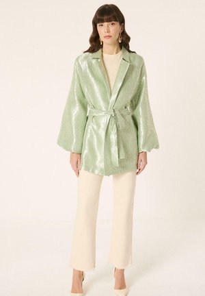 Žena nosi svetlozelenú lesklú bundu s opaskom cez krémový top a krémové široké nohavice, stojí na jednoliatom pozadí.