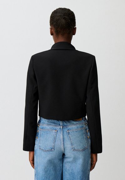 Persona che indossa un blazer nero corto e jeans blu a vita alta con bottoni laterali, di spalle contro uno sfondo chiaro uniforme.