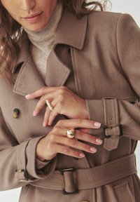 Beige Woll-Trenchcoat mit doppelreihigen Knöpfen, gebundenem Gürtel und strukturierter Oberfläche. Hände zeigen Gold- und Roségoldringe.