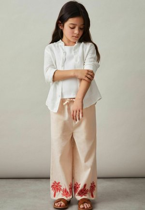 Biele ľanové košeľové tričko s gombíkmi cez biele tielko, doplnené svetloružovými širokými nohavicami so vyšívanými červenými palmami na leme. Hnedé sandále.