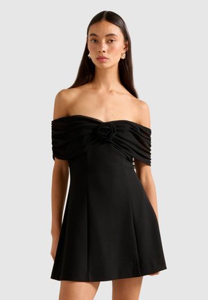 Kvinne som har på seg en svart av-skulderkjole med rynket bodice og rosedetalj i midten, kombinert med gullringøredobber, mot en ensfarget bakgrunn.