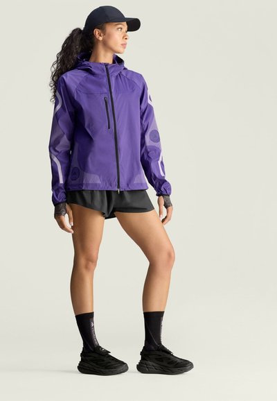 Chaqueta ligera morada con capucha, que presenta mangas de malla y detalles en negro. Combinada con pantalones cortos negros y zapatillas, complementada con calcetines oscuros.