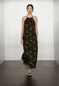 Robe maxi noire à bretelles avec des motifs circulaires dorés, en tissu léger, avec un décolleté froncé et des fentes sur les côtés ; portée avec des sandales noires.