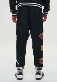 Zwarte joggings met gestreepte boorden, voorzien van witte tekst en teamlogo's in rood en goud. Zachte stof met een ontspannen pasvorm.