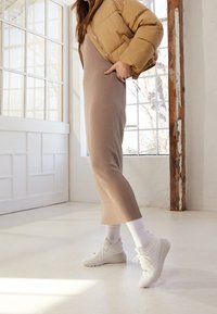 Beige ribbed knit dress, mid-calf length, paired with a tan puffer jacket and white athletic shoes, with a minimalistic interior backdrop.