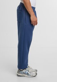 Blauwe trainingsbroek met elastische tailleband, zijzak en een gladde textuur; gecombineerd met zilverkleurige hardloopschoenen met blauwe accenten.