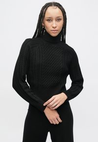 Crni pleteni džemper s vratom na preklop s teksturiranim uzorkom, rebrastim manšetama i rubom te krojem koji prianja uz tijelo. Nošen uz glatku crnu odjeću.