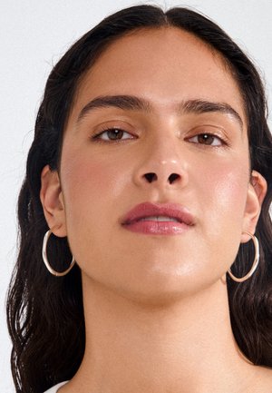 Nainen, jolla on tumma laineikas hiukset, käyttää suuria hopeisia rengaskorvakoruja, luonnollista meikkiä ja neutraalia huuliväriä, katsoo hieman ylöspäin vaaleaa taustaa vasten.