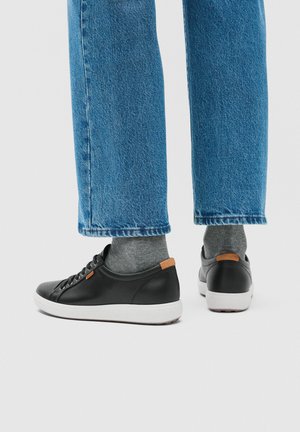 Schwarze Sneakers mit niedrigem Schaft, weißen Sohlen und braunen Fersenlaschen, getragen mit grauen Socken und blauen Jeans vor einem schlichten hellen Hintergrund.
