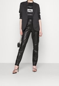 Blazer negro de rayas, pantalones de cuero negros ajustados, tacones negros con tiras. La modelo sostiene un pequeño bolso negro con una correa de cadena.