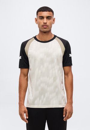 Man met kort donker haar en snor, draagt een beige en zwart sportshirt met korte mouwen en een logo op de mouwen, staand tegen een effen achtergrond.