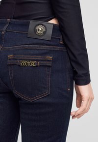 Κοντινή λήψη από σκούρο μπλε τζιν Versace Jeans Couture με χρυσό λογότυπο στην τσέπη και μαύρη δερμάτινη ετικέτα στην ταινία μέσης.