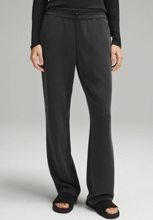 Osoba nosząca luźne czarne spodnie ze ściągaczem w pasie, w połączeniu z czarnymi sandałami typu slide i czarną bluzką z długim rękawem, stojąca na jednolitym tle.