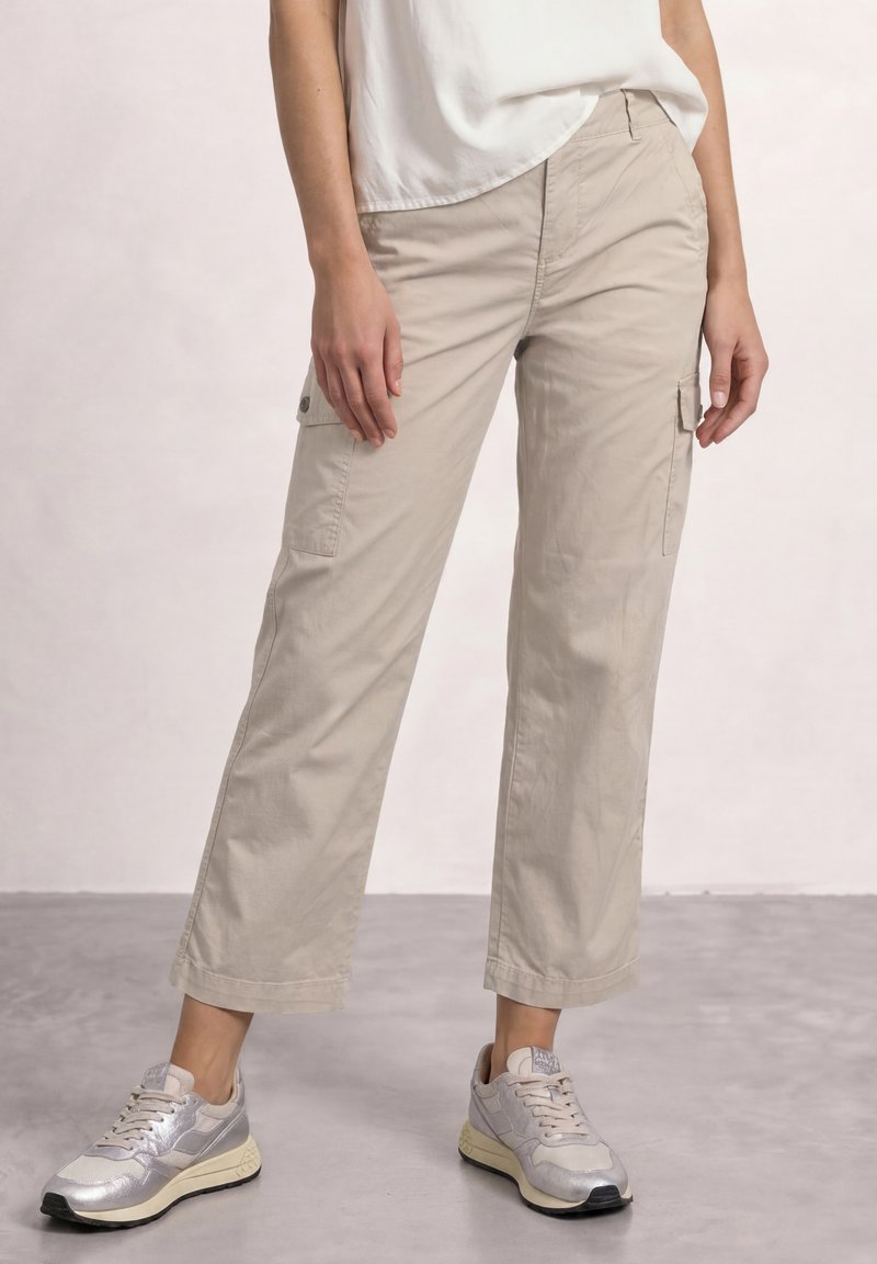 Personne portant un pantalon cargo beige à jambes larges, un haut blanc et des baskets argentées, debout sur un sol gris avec un fond clair.
