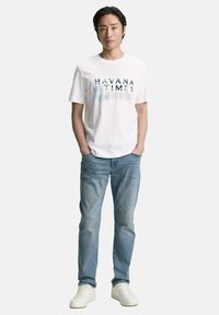 Camiseta blanca con gráfico de "HAVANA TIMES", combinada con jeans de mezclilla azul claro y zapatillas blancas. Ajuste casual, mangas cortas, textura suave.