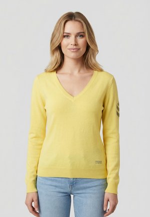 Woman with blonde hair wearing a yellow V-neck sweater and light blue jeans, standing against a plain light background.