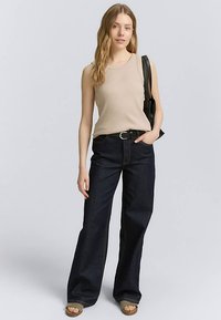 Haut beige sans manches associé à un jean ample bleu foncé, complété par une ceinture noire et un sac à bandoulière noir. Sandales marron clair.