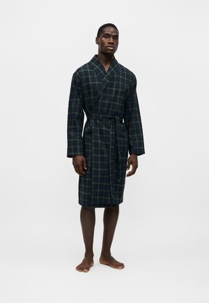 Homme debout pieds nus portant un peignoir à carreaux vert foncé et bleu marine jusqu'au genou, noué à la taille, sur un fond blanc uni.