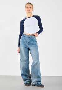 Junge Frau steht vor einem weißen Hintergrund und trägt ein weißes und marineblaues Langarm-Crop-Top, weit geschnittene Jeans und marineblaue Sneakers.
