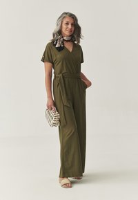 Olijfgroene wikkeloverall met korte mouwen, zelfbindende riem, wijde benen en zakken. Gecombineerd met een gestreepte clutch en slide sandalen.