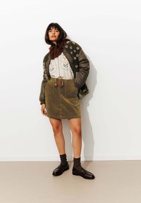 Femme portant un chemisier brodé de fleurs, une jupe en velours côtelé vert olive, une veste florale marron, une écharpe en tricot, des chaussettes noires et des mocassins marron, se tenant devant un mur blanc.