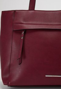 Sac cabas en cuir bordeaux avec poche avant zippée, deux poignées supérieures et plaque de marque argentée en bas à droite.