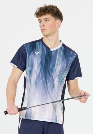 Ung mand holder en badmintonketcher, iført en blå- og hvidmønstret sportstrøje og marineblå shorts, og kigger til siden.
