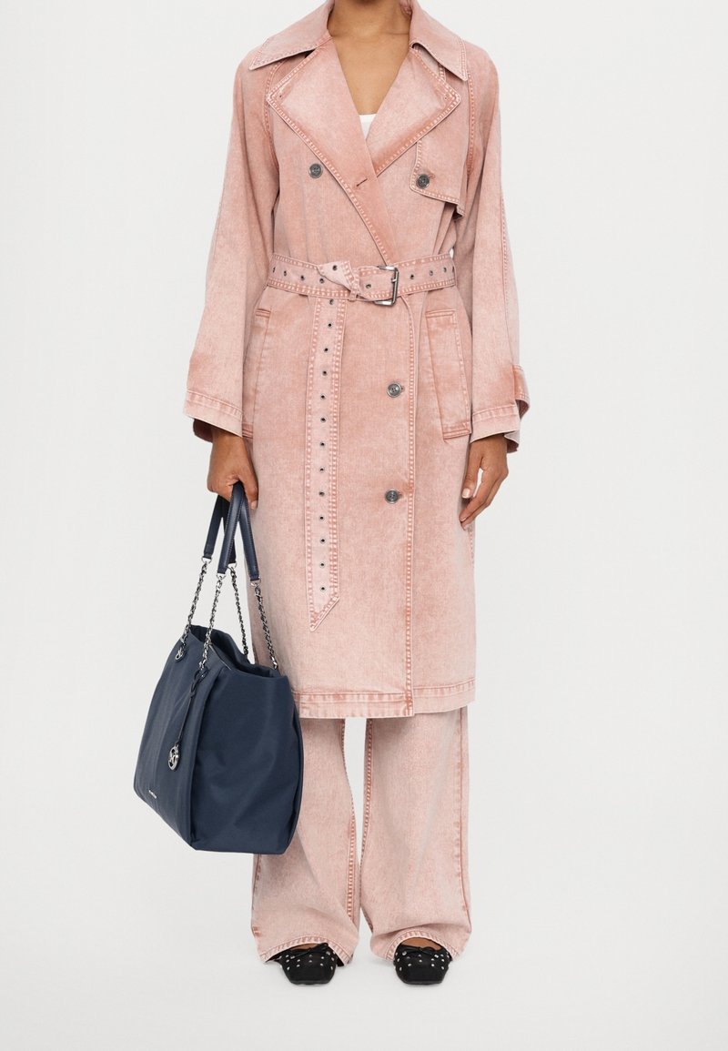 Personne portant un trench-coat ceinturé rose clair et un pantalon large assorti, tenant un grand sac à main bleu marine avec des anses en chaîne.