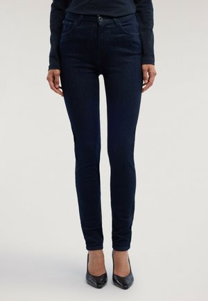 Kvinna som bär mörkblå slim-fit jeans och svarta högklackade skor, står mot en slät ljus bakgrund.