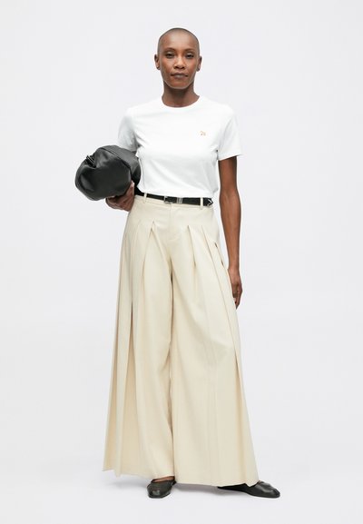 Žena s oholenou hlavou, oblečená v bílé tričko, béžových volných skládacích kalhotách, s černým páskem a botami, držící černou koženou tašku na bílém pozadí.