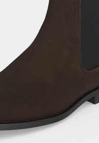Gros plan d'une botte cheville en daim marron foncé avec un panneau latéral élastique noir et une semelle noire sur un fond clair.