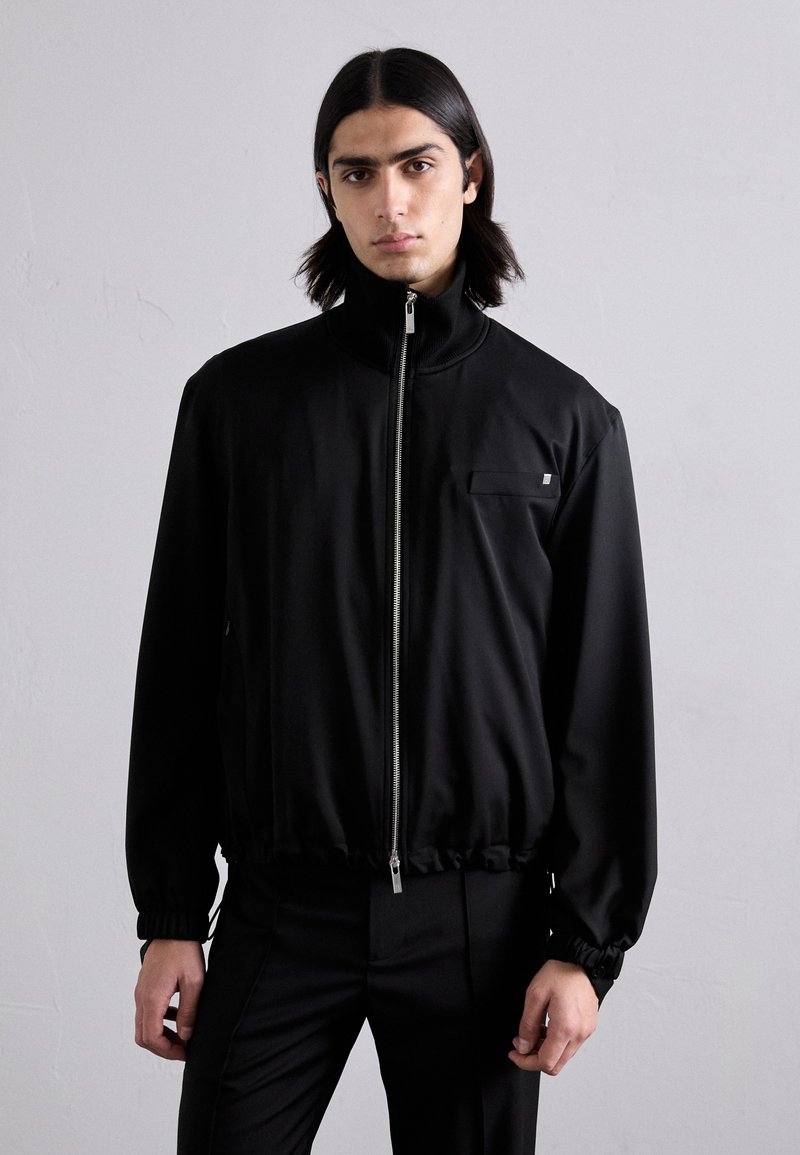 Black windbreaker jacket with a high collar, front zipper, and elastic cuffs. Features a small chest pocket and a smooth, lightweight fabric.