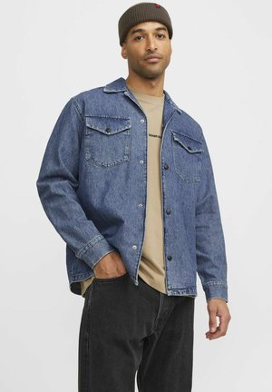 Denim jacket in medium blue with two chest pockets, button closure, and casual fit. Worn over a beige t-shirt and dark pants. Brown knit beanie.