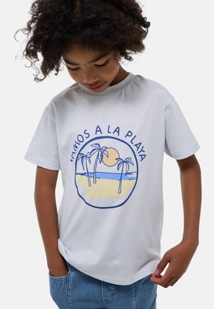 T-shirt en coton blanc avec un imprimé graphique de palmiers et d'un soleil sur un paysage de plage en bleu et jaune, col rond, manches courtes.