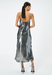 Metallisch graues Slipkleid mit dünnen Trägern, offenem Rücken und fließender Textur, kombiniert mit schwarzen High-Heelsandalen und Schnürdetails.