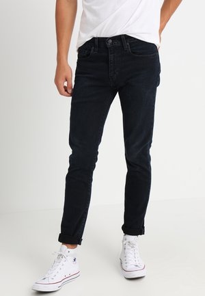 Man met opgerolde zwarte jeans, witte hoge sneakers en een wit T-shirt, staand tegen een effen achtergrond.