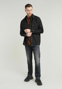 Zwart denim overhemdjasje met twee voorzakken, gedragen over een bruine poloshirt. Grijze jeans en zwarte sportschoenen maken de outfit compleet.