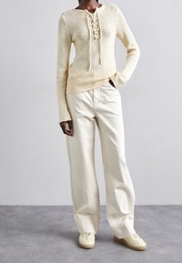 Pull en tricot jaune clair avec un devant à lacets, associé à un pantalon évasé crème et des chaussures beiges. Tissu texturé, design décontracté.