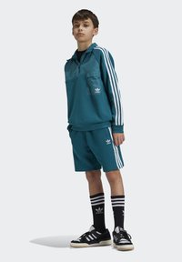 Conjunto deportivo en verde azulado con una parte superior con cremallera y bloques de color, pantalones cortos a juego, calcetines negros con rayas blancas y zapatillas negras con tres rayas.