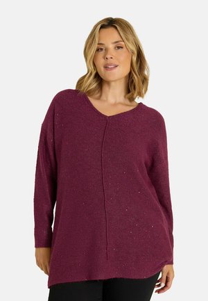 Femme aux cheveux blonds mi-longs portant un pull ample à manches longues violet foncé et un pantalon noir sur fond blanc.