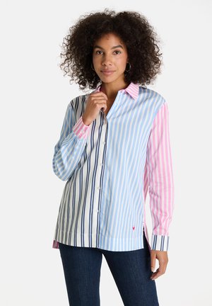 Frau mit lockigem Haar, die ein langärmliges Hemd mit vertikalen Streifen in Marineblau, Blau, Weiß und Pink trägt, kombiniert mit dunkler Jeans.