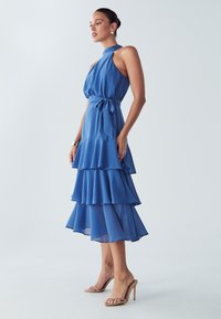 Ženska stoji v modri obleki s tkaninami in vratnim trakom, zavezanim okoli pasu, obuta v bež sandale z visokimi petami in zlato nakit.