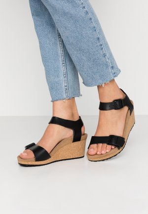 Sorte kile-sandaler med korkbund; udstyret med to stropper, en justerbar spænde og en glat overflade. Kombineret med slidte lyseblå jeans.