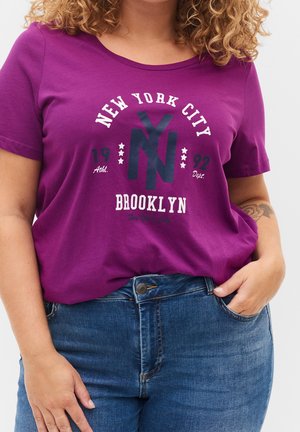 Personne portant un t-shirt violet avec le texte "New York City Brooklyn 1992" et un jean bleu, main dans la poche, tatouage visible sur le bras.