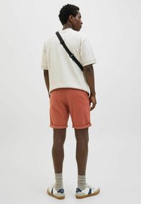 Men's outfit featuring a beige oversized t-shirt, rolled-cuff coral shorts, gray socks, and white sneakers with navy accents. Black shoulder strap visible.