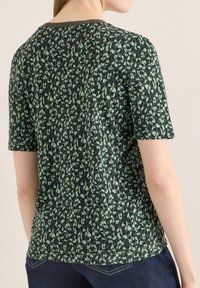 Femme portant un haut à manches courtes avec un imprimé léopard abstrait vert et noir, associé à un jean bleu foncé, vue de dos contre un mur beige.