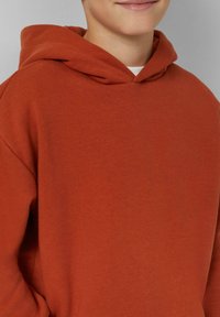 Hoodie de couleur rouille en coton doux, avec une capuche à lien de serrage et une coupe décontractée. Pas de logos ni de motifs visibles.